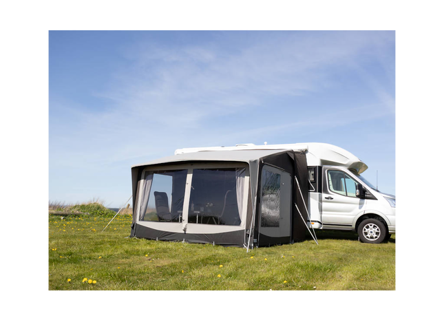 Camping trailer with an extended awning on a grassy field under a blue sky.