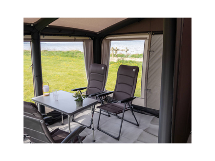 Camping trailer interior with chairs and a table near large windows showing greenery outside.