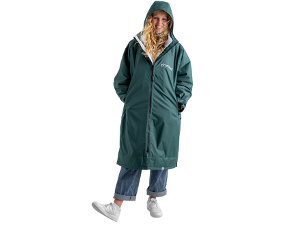 A woman wearing a long green hooded dryrobe with a white inner lining, blue jeans, and white sneakers, standing against a plain white background.