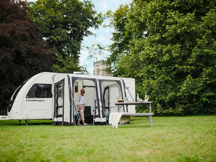 Caravan with an awning on a grassy area with trees and a castle in the background