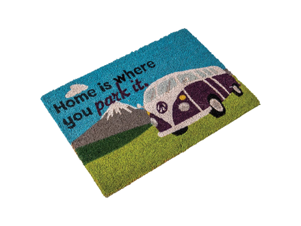 A rectangular coir doormat with a colorful printed design featuring the text 'Home is where you park it.' alongside a purple and white vintage camper van on a green grassy field under a blue sky with a mountain in the background.