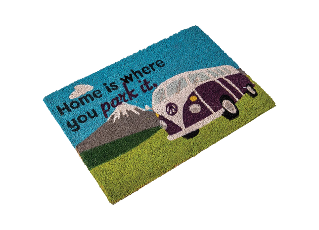 A rectangular coir doormat with a colorful printed design featuring the text 'Home is where you park it.' alongside a purple and white vintage camper van on a green grassy field under a blue sky with a mountain in the background.