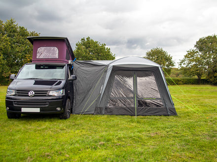 Van with a large gray awning extended on a grassy field with trees in the background