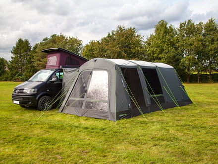 Van with a large gray awning extended on a grassy field with trees in the background