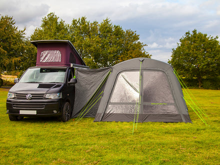 Van with a roof tent and an attached air awning on a grassy field with trees in the background.
