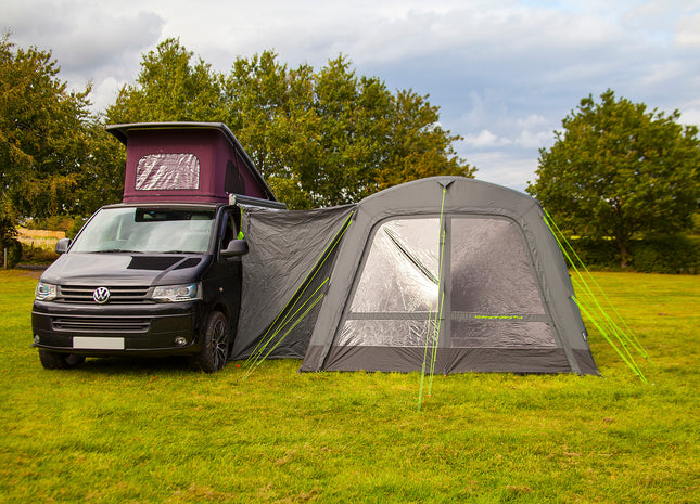 Van with a roof tent and an attached air awning on a grassy field with trees in the background.