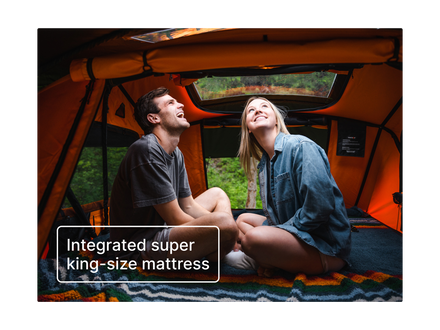 A man and a woman sitting inside a tent, smiling and looking up at the integrated roof window. The tent interior is orange, and a text overlay at the bottom reads 'Integrated super king-size mattress'.