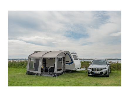 Caravan with an attached awning and a car parked nearby on a grassy area with a cloudy sky.