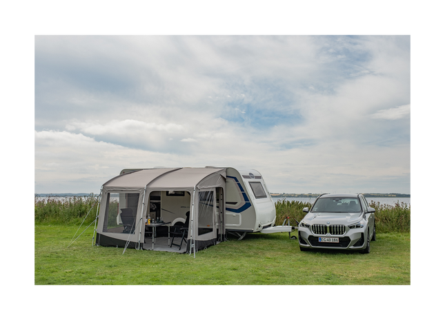 Caravan with an attached awning and a car parked nearby on a grassy area with a cloudy sky.
