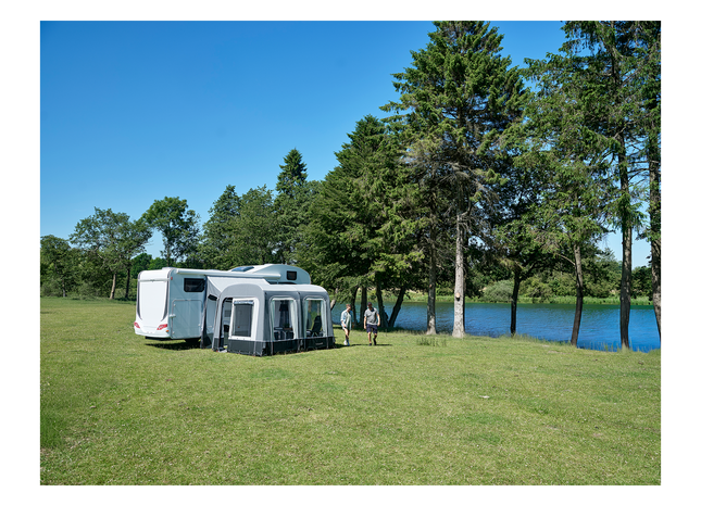 caravan with attached awning on a grassy area near a body of water with trees in the background.