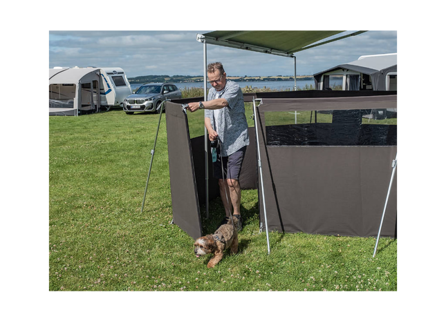 Man setting up a camping awning with a dog in a grassy area with other campers in the background.